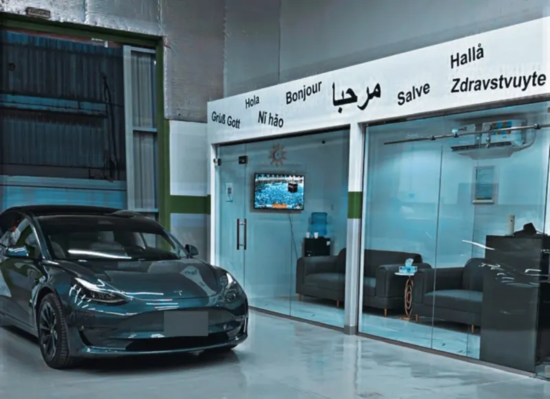 A dark grey Tesla Model 3 parked outside the glass-walled customer lounge at EV Zone, under a sign that says hello in several languages