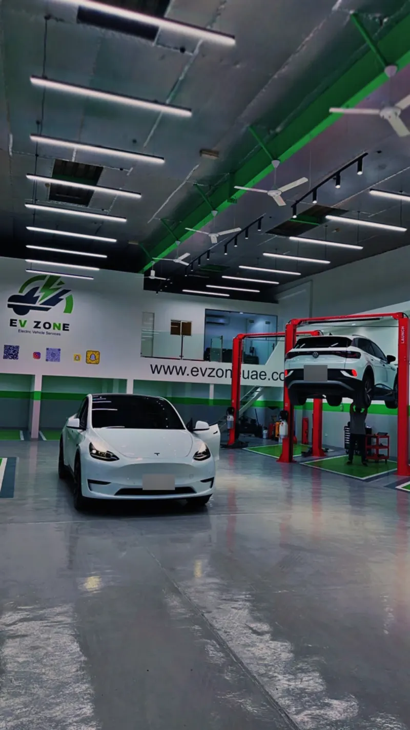 The EV Zone workshop floor: a white Tesla Model Y in front, and a white VW ID.4 up on a red two-post lift with a technician working underneath